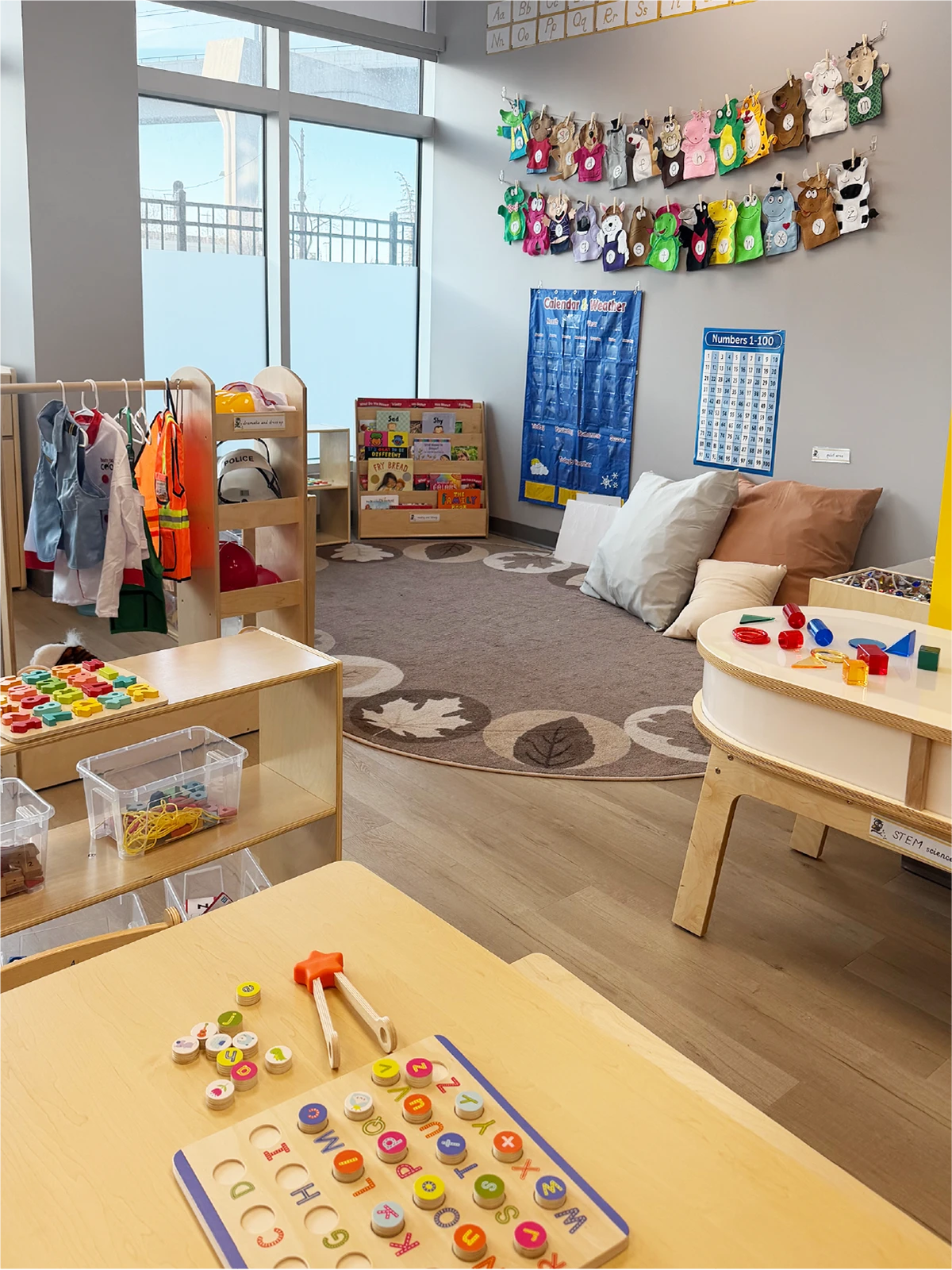 A bright, organized preschool classroom in one of the best early learning centres, with toys on tables, a rug with leaf patterns, dress-up clothes, colorful crafts on the wall, and an activity calendar in the corner.