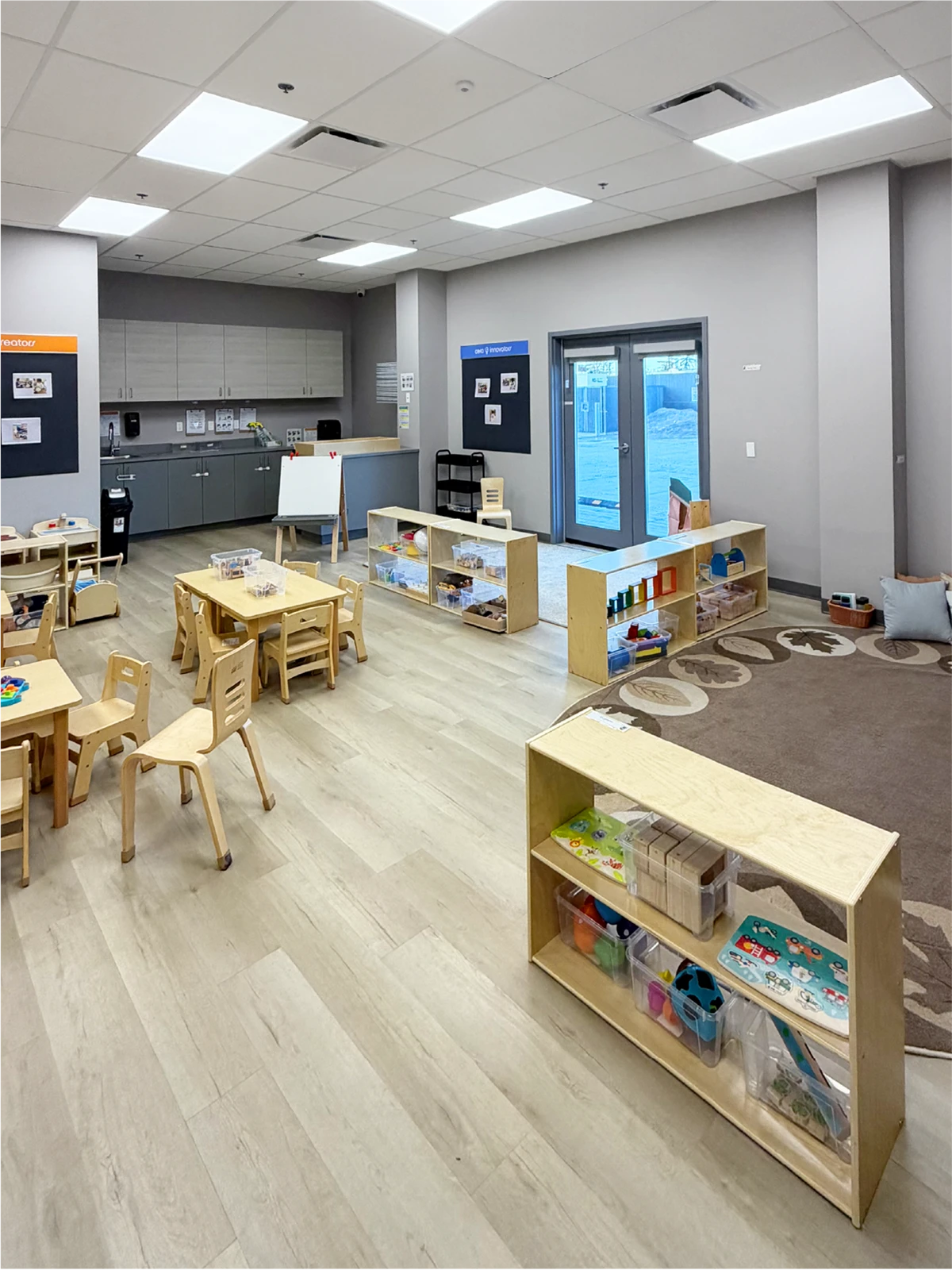 A bright, organized early learning centre classroom with wooden tables and chairs, low shelves holding educational toys and books, a rug area, and large windows letting in natural light.