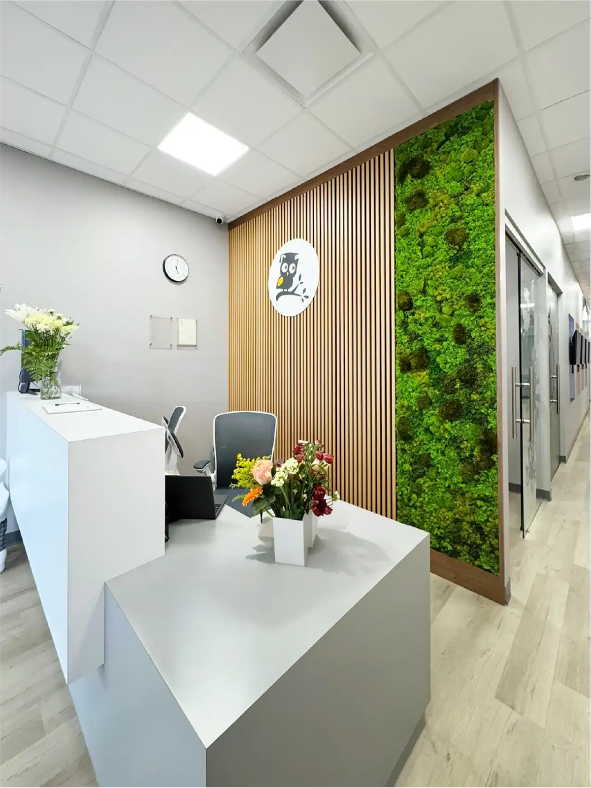 A modern reception area for early learning centres features a white desk, a chair, and two vases of flowers. Behind the desk is a wooden slat wall with an owl logo, a vertical green moss wall, and a hallway with glass doors to the right.
