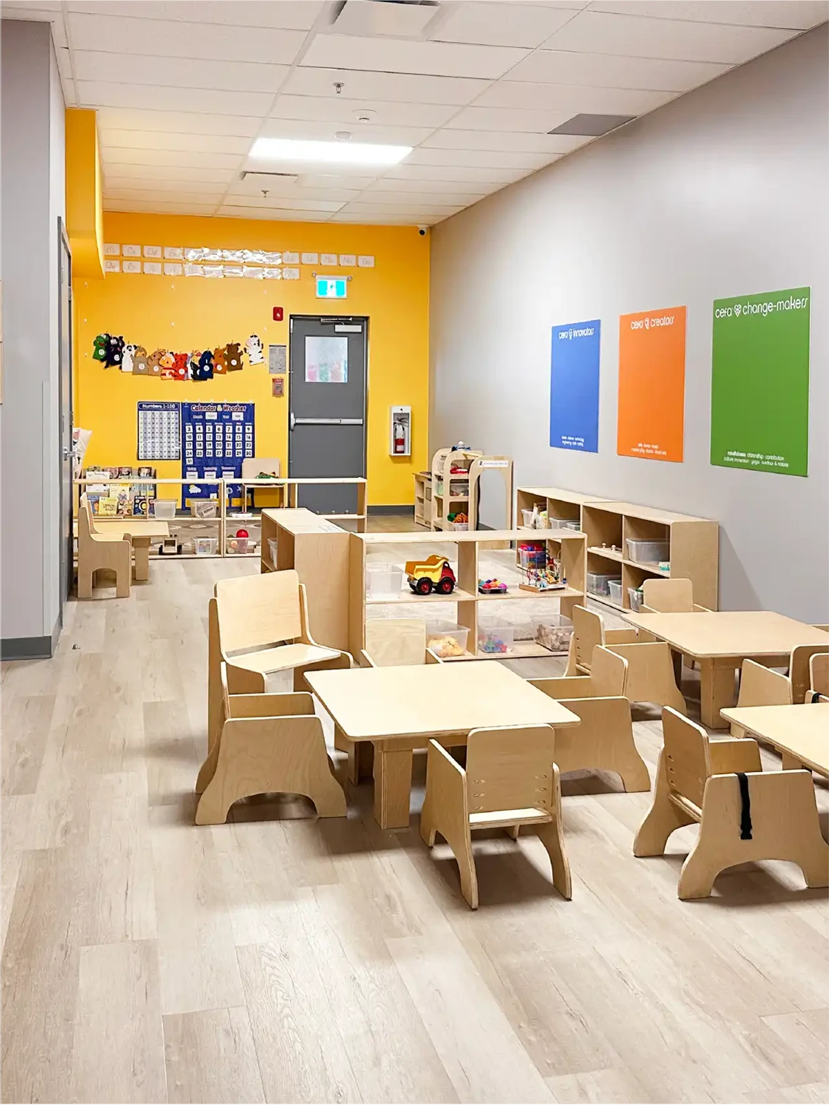 A brightly lit preschool classroom at one of the leading early learning centres, with light wood tables and chairs, colorful wall decorations, organized shelves with toys and books, a yellow accent wall, and a clean, welcoming atmosphere.