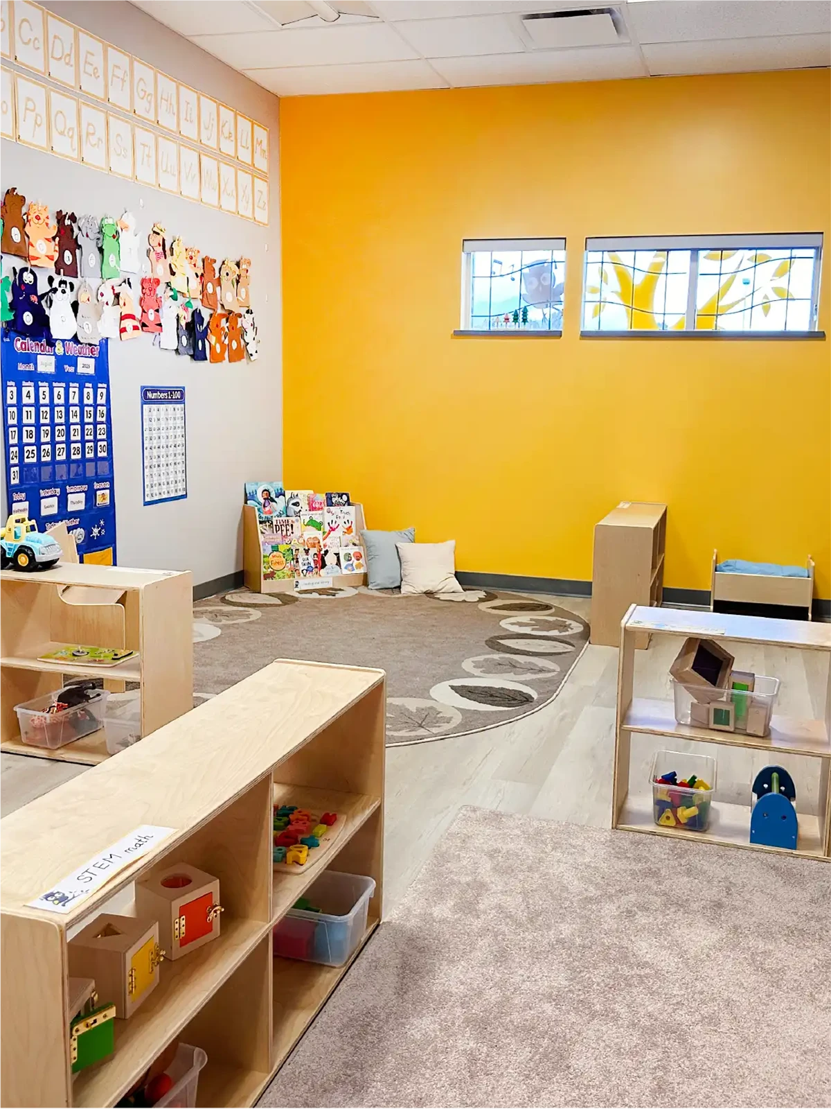 A brightly lit preschool classroom with yellow walls, low wooden shelves holding toys, a rug with cushions, educational posters, and artwork. Two windows let in natural light—perfect for families searching for preschools close to me or quality early learning centres.