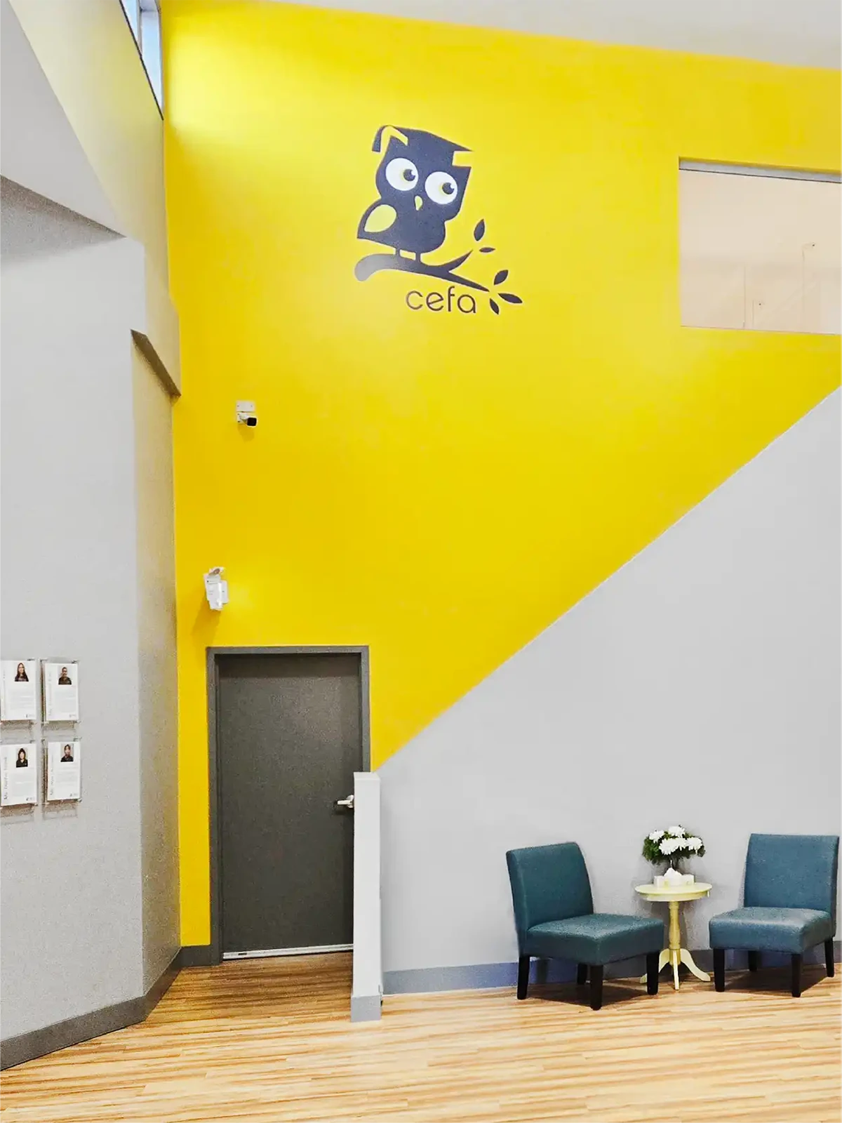 A modern interior with a yellow and gray wall, featuring a cartoon owl and the word "cefa," evokes the playful spirit of early learning centres. Below are two teal chairs, a small round table with a flower vase, and a dark gray door.