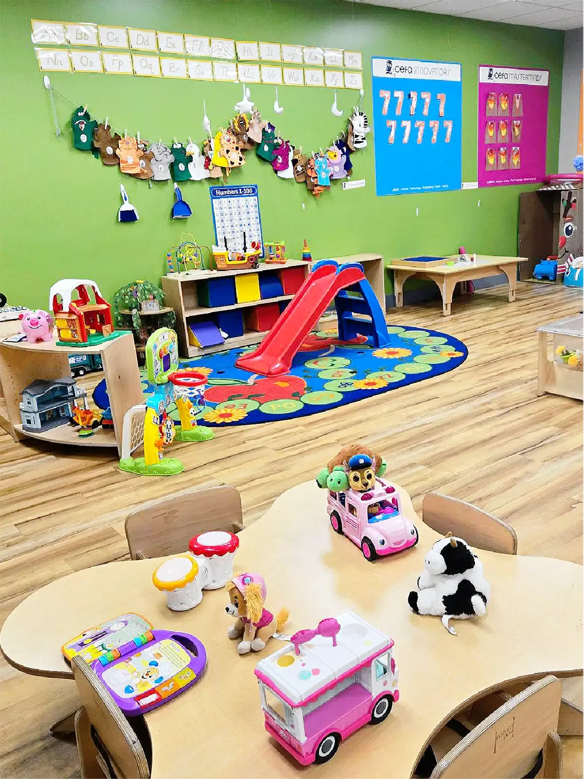 Colorful preschool classroom in one of the top early learning centres, with wooden floors, a play area with a red slide, alphabet and number posters on the walls, and small tables filled with toy cars, stuffed animals, and learning games.