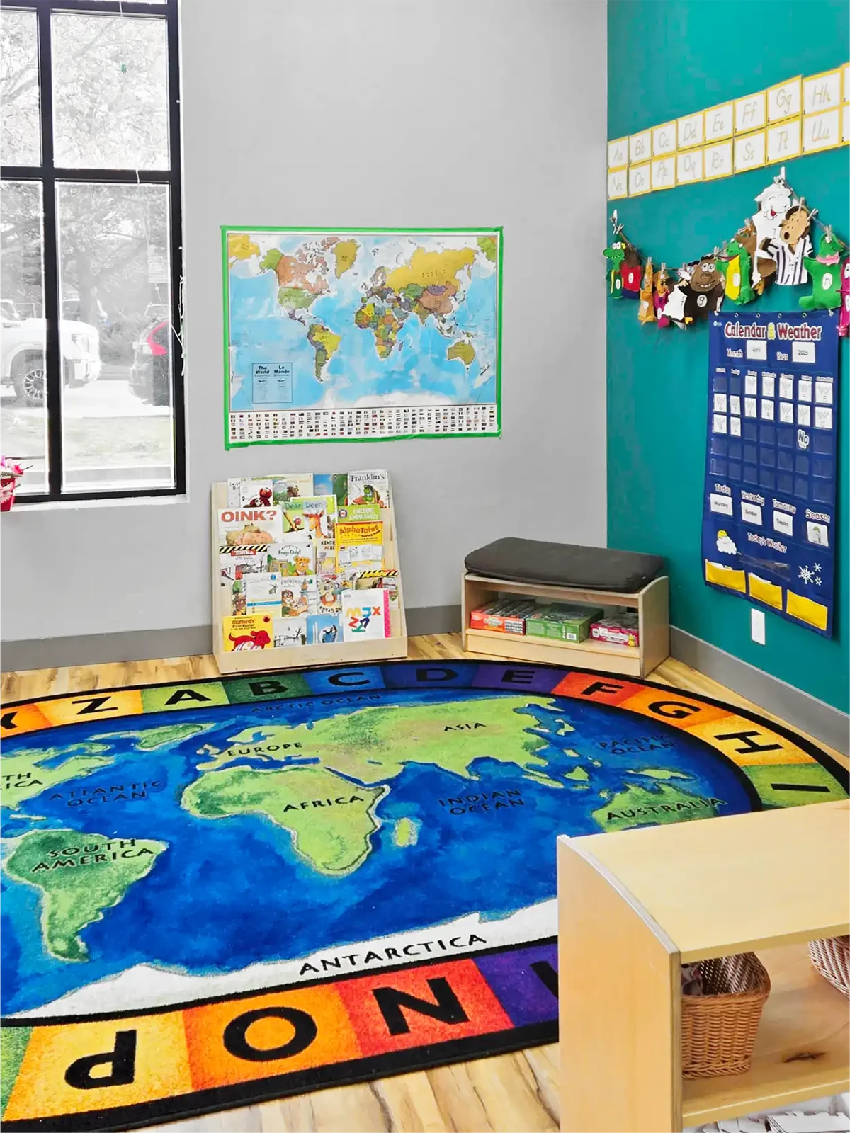 A bright classroom corner at one of the top early learning centres, featuring a world map rug, alphabet border, books in a rack, a calendar chart, and colorful decorations on a teal wall. A large window fills the space with natural light.