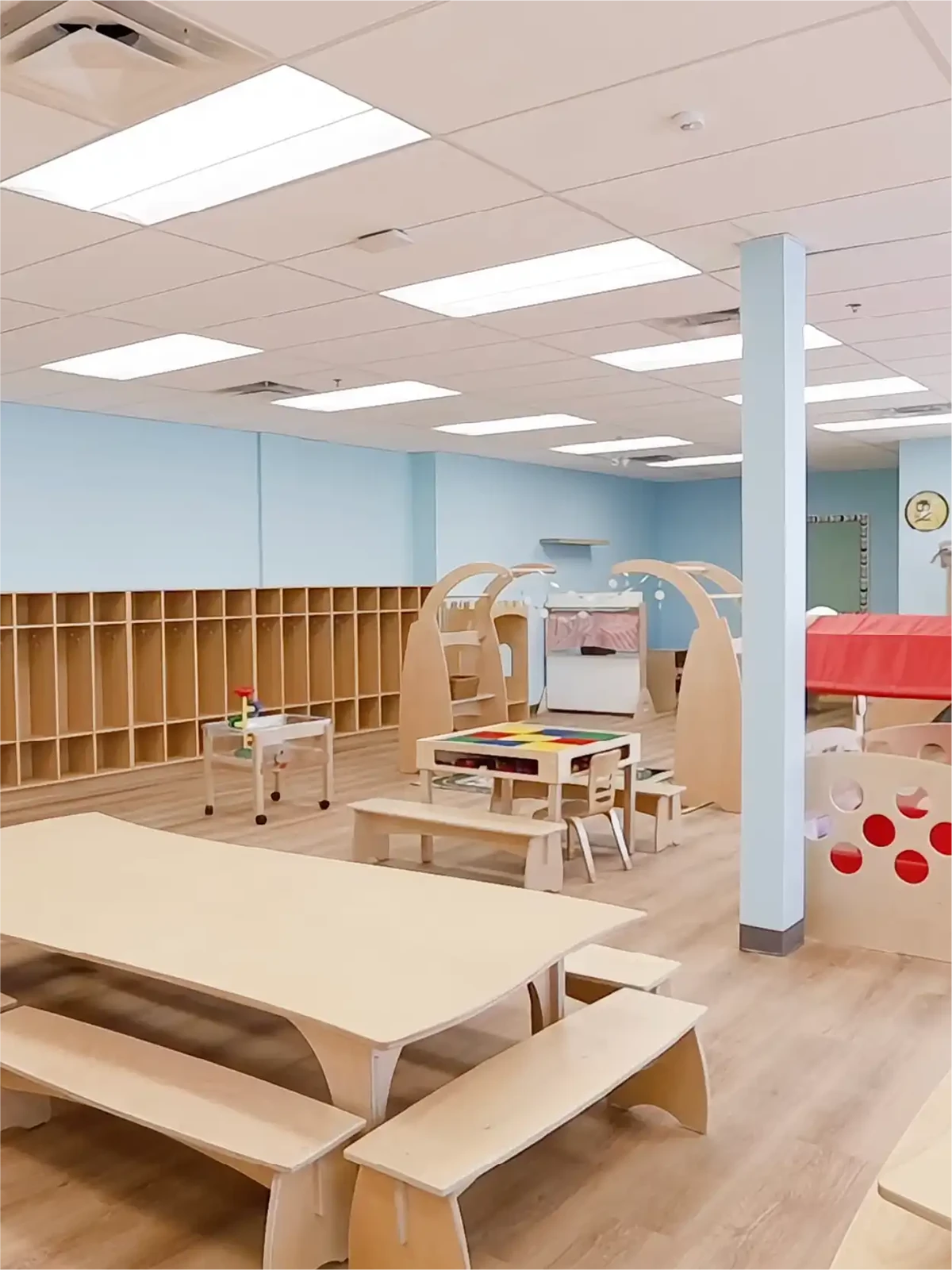 A bright, spacious classroom in one of the top early learning centres, featuring light wood tables, benches, cubby shelves, play structures, and toys. Light blue walls and large overhead lights complement the wood-patterned floor.