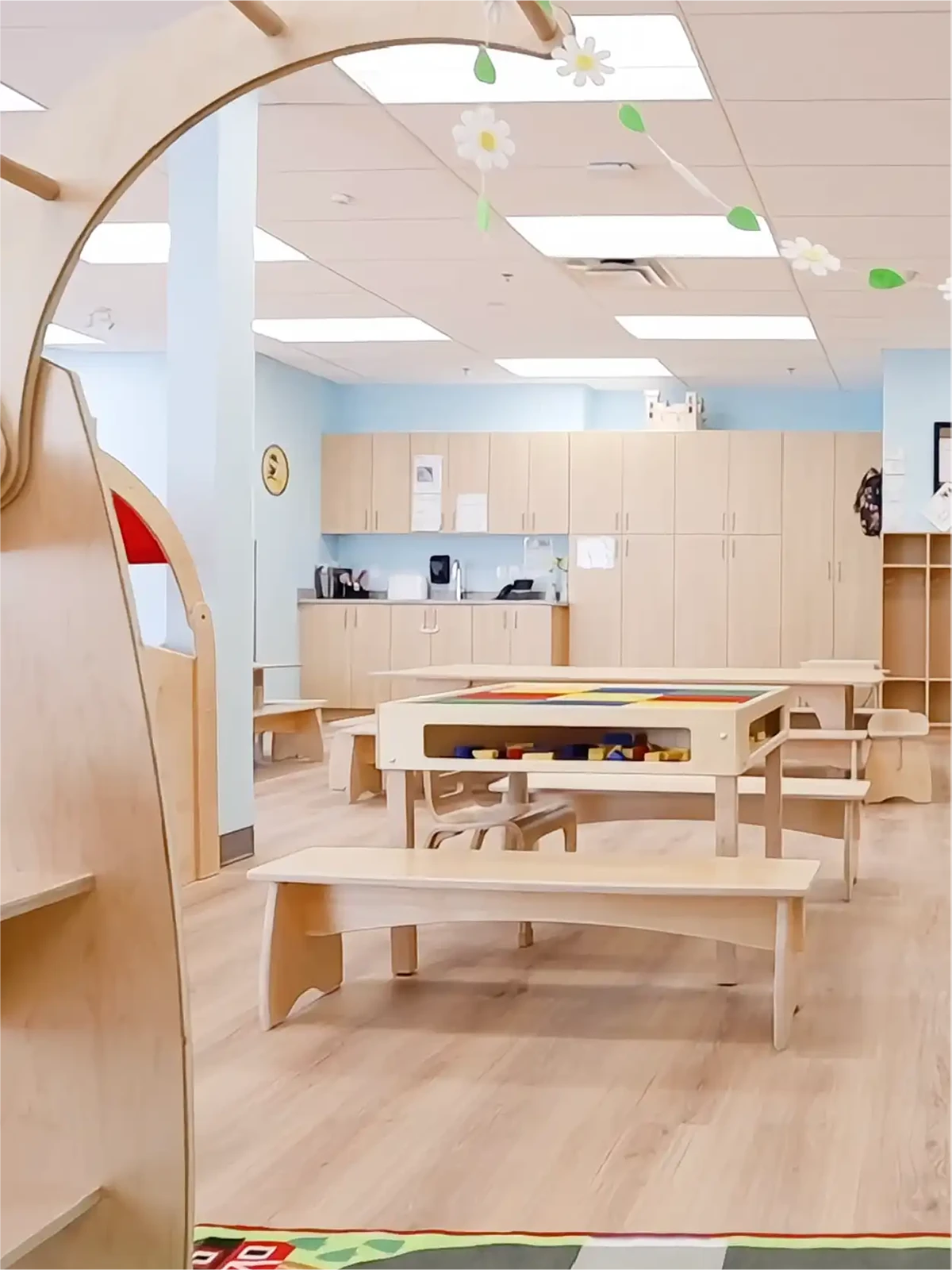 A bright, tidy classroom in one of the best early learning centres features child-sized tables, play equipment, and light blue walls. Decorative flowers hang from the ceiling, making it an organized and inviting space for children.