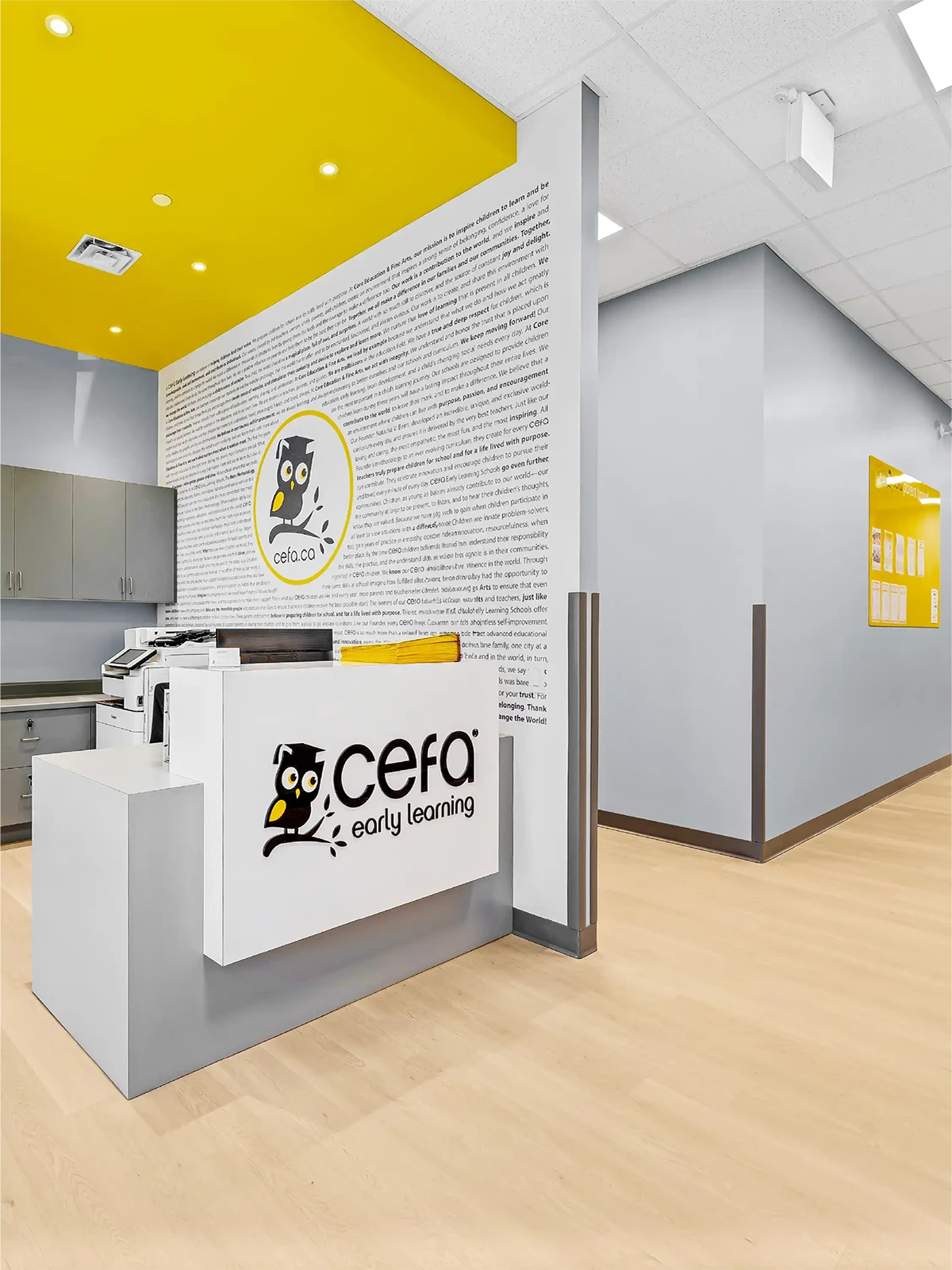 A modern, brightly lit reception area at CEFA Early Learning, one of the leading early learning centres, featuring light wood floors, grey walls, yellow accents, and a white front desk with the CEFA logo and an owl graphic.