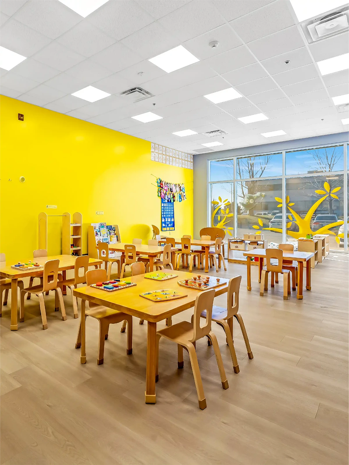 A bright classroom in early learning centres with yellow walls, wooden tables and chairs, educational materials on the tables, and large windows decorated with yellow designs letting in natural light.