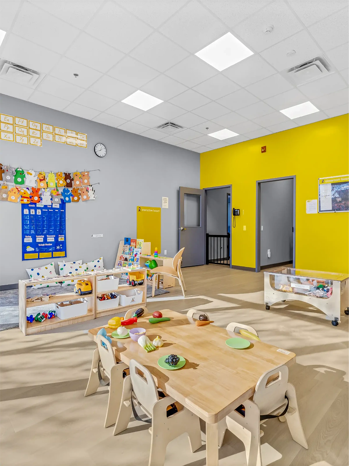 A bright, colorful preschool classroom with yellow and gray walls, small wooden tables and chairs, play food, toys, shelves with supplies, and educational posters—perfect for centres for early learning.
