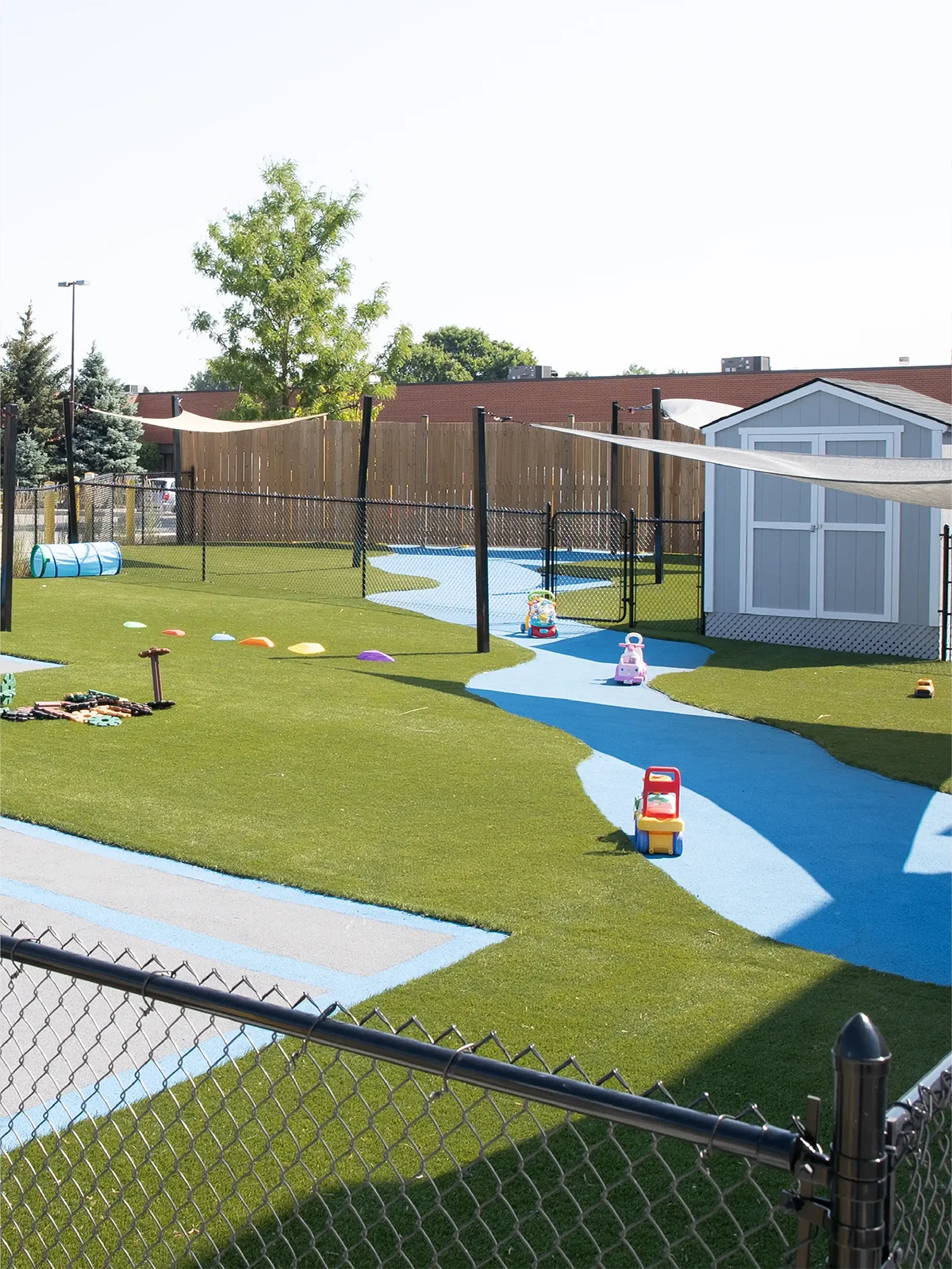 An outdoor playground at one of the top early learning centres, featuring artificial grass, a winding blue path, toy cars, play equipment, and a white shed within a black fence. Trees and nearby buildings provide a welcoming backdrop.