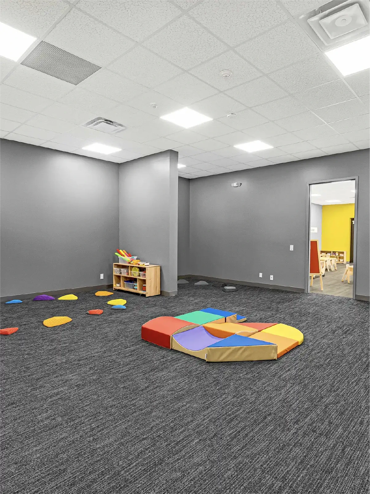 A spacious, gray-walled playroom—found in many early learning centres—features carpeted floors, colorful foam mats, stepping stones, and a small wooden shelf with toys. An open doorway leads to a bright yellow room in the background.
