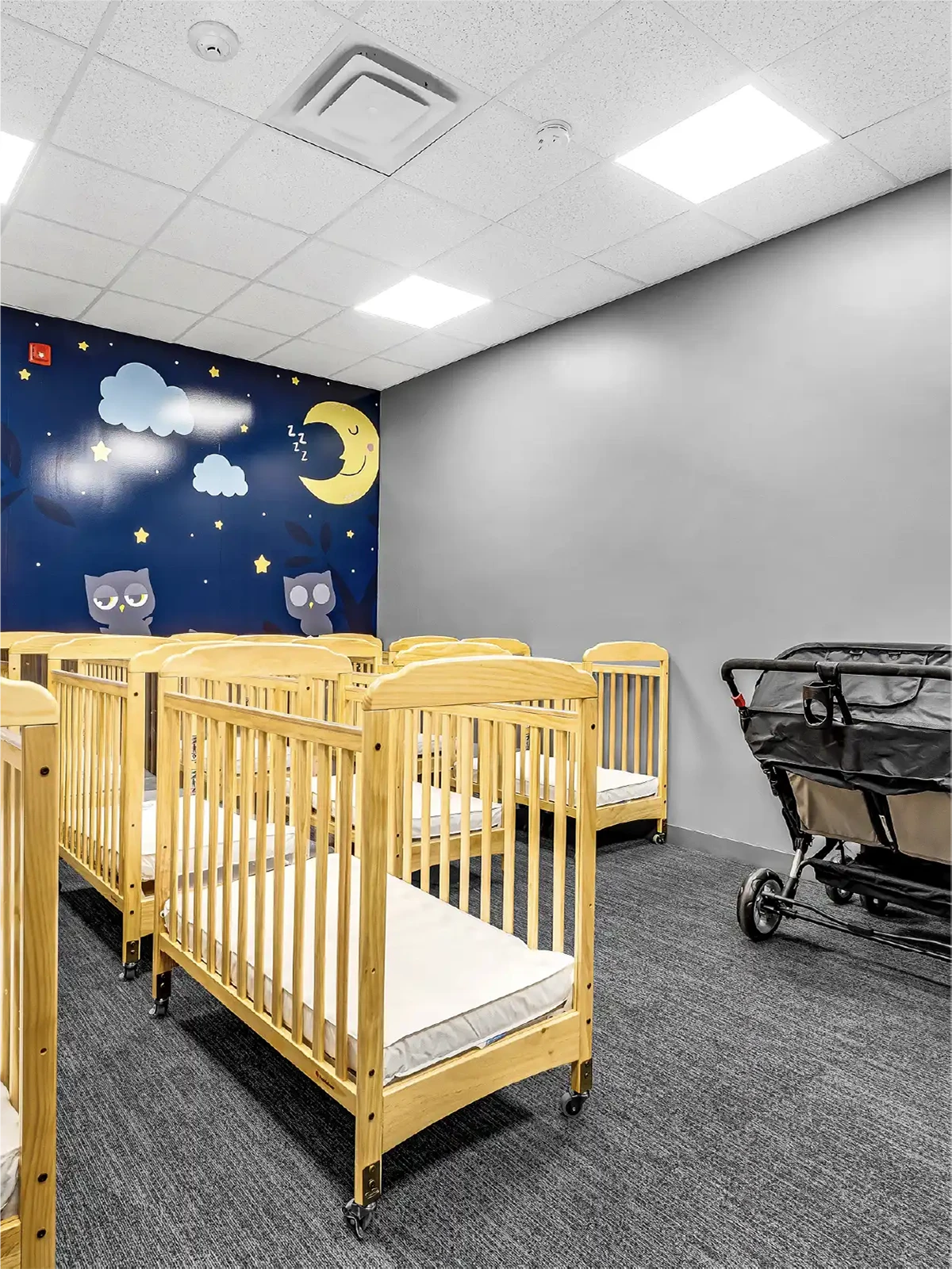A brightly lit nursery at one of the leading early learning centres, featuring empty wooden cribs, a large stroller, and a whimsical wall mural with owls, clouds, stars, and a moon on a dark blue background—perfect for little learners.