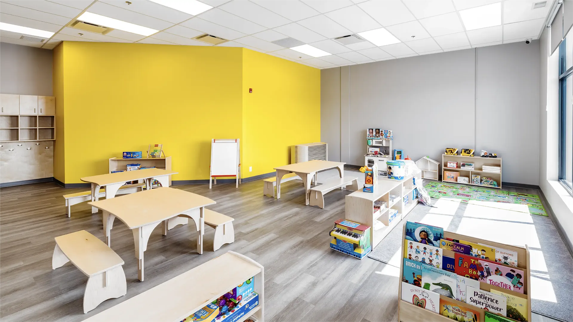 Bright classroom with yellow and gray walls, wooden tables and benches, bookshelves filled with children's books—perfect for early learning centres—with an easel, play area, and colorful rug near large windows.