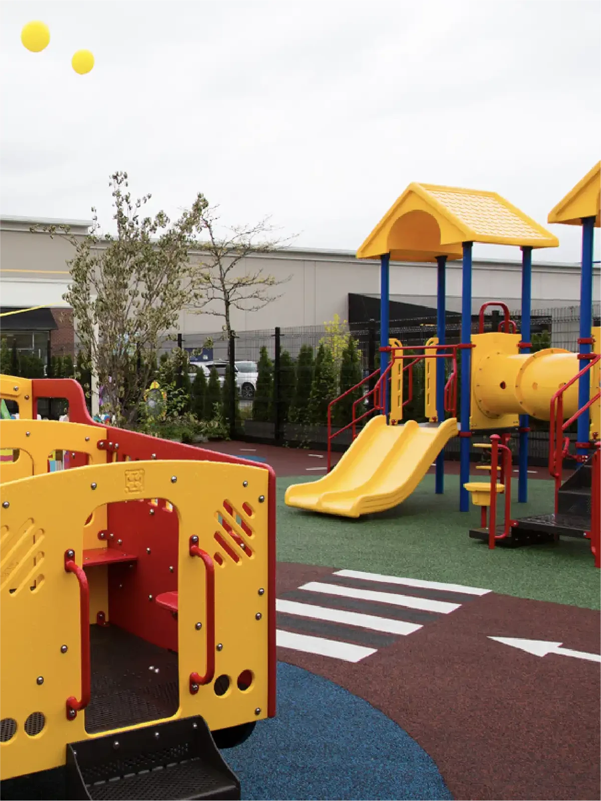 Colorful playground with yellow and red play structures, slides, climbing equipment, and a tunnel—perfect for early learning centres. Surrounded by trees and a fence, two yellow balls float in the sky.