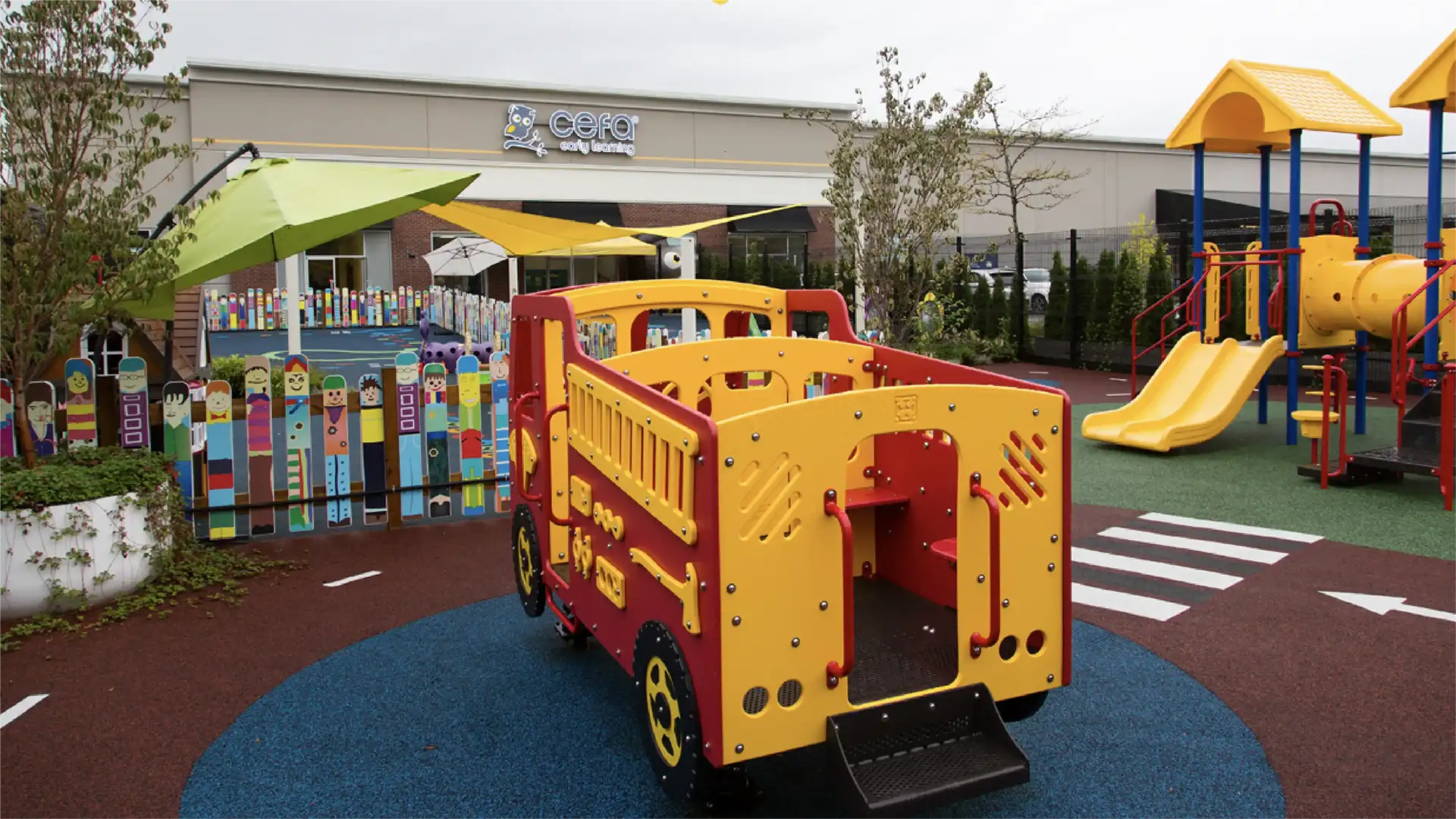 Colorful outdoor playground at one of the top early learning centres, featuring a large red and yellow play vehicle, slides, climbing equipment, artificial turf, and a fence with painted pickets. A daycare or preschool schools near me building is in the background.