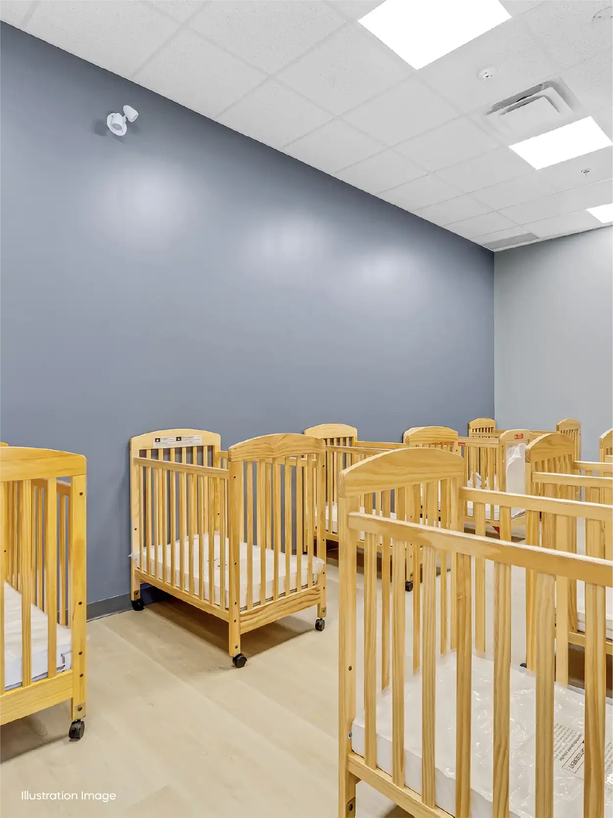 A bright nursery room in one of the top early learning centres, featuring a row of empty wooden cribs along the walls, light wood flooring, a blue accent wall, and ceiling lights above.