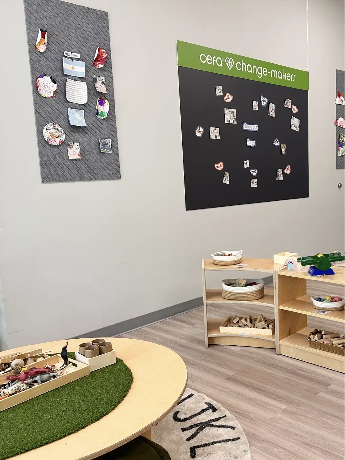 A classroom corner in one of the early learning centres with low wooden shelves holding educational toys and materials, a round table with artificial grass, and a wall display labeled "cefa change-makers" featuring children's artwork.