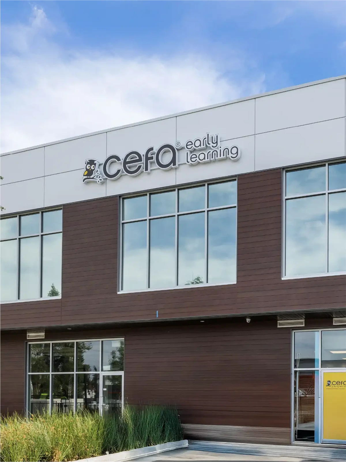 A modern building with large windows and a sign that reads "cefa early learning," featuring a small owl logo above the entrance. Green plants sit out front, making it a welcoming choice among preschool schools near me under a blue sky.