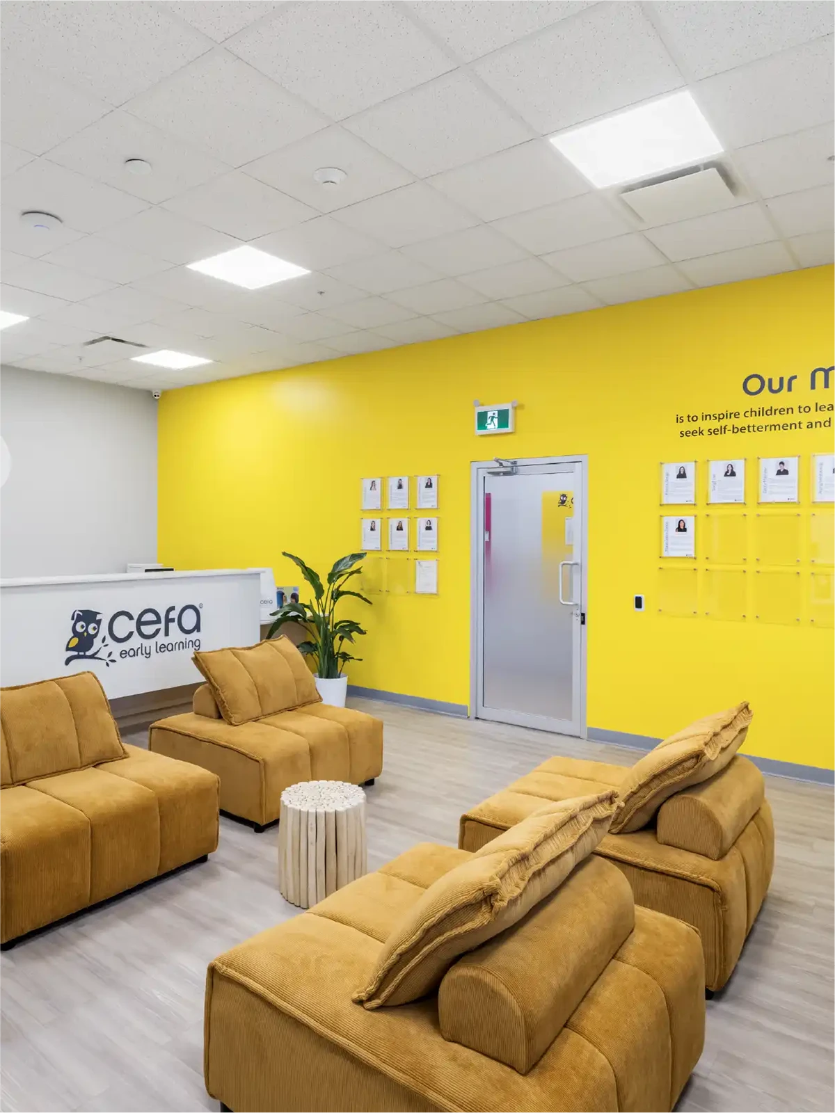 A bright, modern waiting area with mustard yellow sofas, wooden tables, and a vibrant yellow accent wall featuring inspirational text and framed photos welcomes families to one of the top early learning centres in your area.