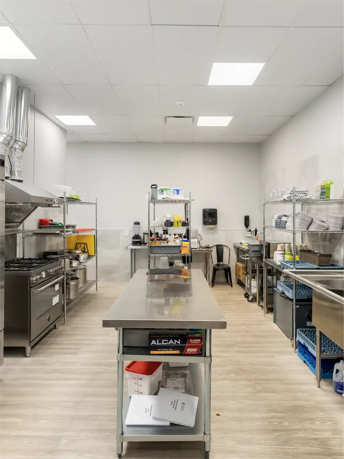 A clean, modern commercial kitchen with stainless steel appliances and countertops, metal shelving stocked with dishes and supplies, and bright overhead lighting—ideal for centres for early learning or preschool schools near me.