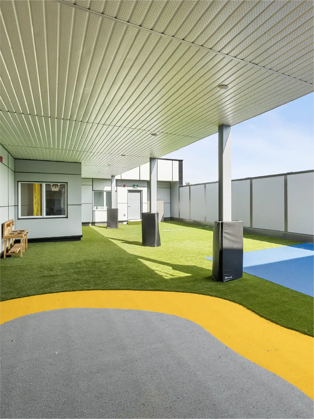 A covered outdoor play area at early learning centres, featuring artificial grass, colorful rubber flooring in gray, yellow, and blue, benches, padded columns, and a fenced perimeter under a white ceiling.