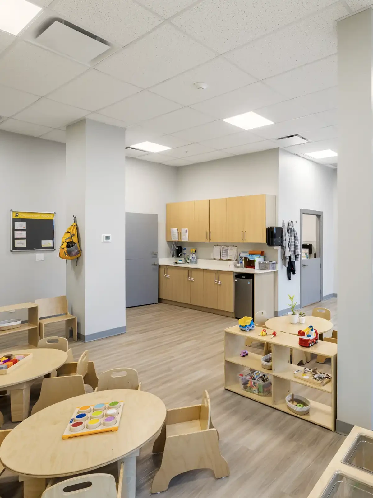 A bright, modern daycare classroom at one of the local early learning centres, featuring small wooden tables and chairs, open toy shelves, cabinets, a bulletin board, coat hooks, and a backpack on the wall with light wood floors and white walls.