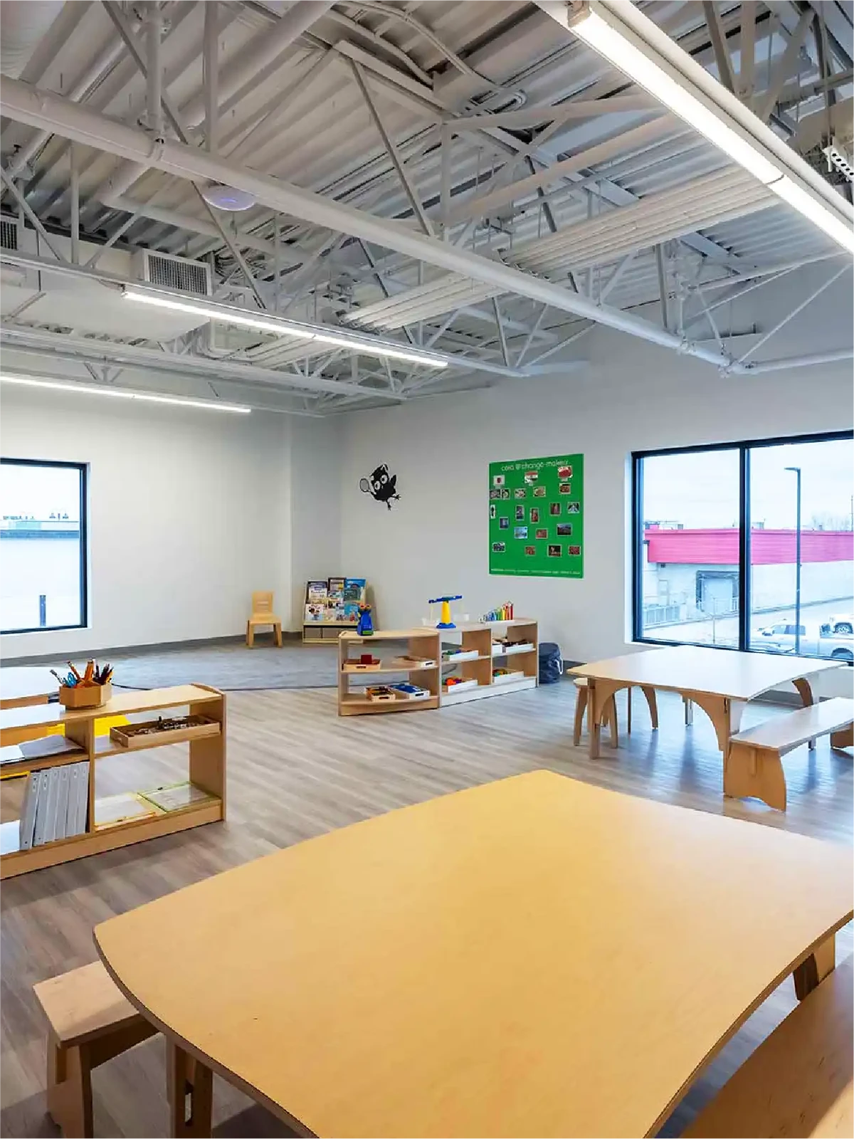 A bright, spacious classroom in early learning centres with large windows, light wood tables and shelves, art supplies, toys, and a green bulletin board. High ceilings with exposed beams fill the room with natural light from outside.