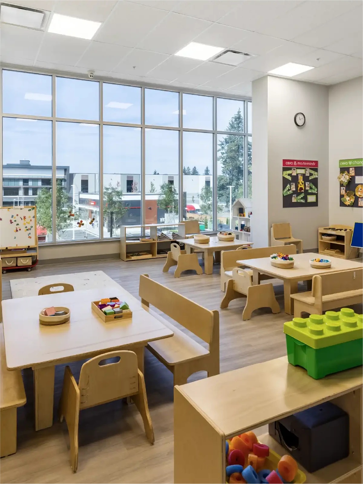 Bright, modern classroom in one of the leading early learning centres with large windows, wooden tables and chairs, educational toys, a whiteboard, and colorful posters. Outdoor buildings and trees are visible through the windows.