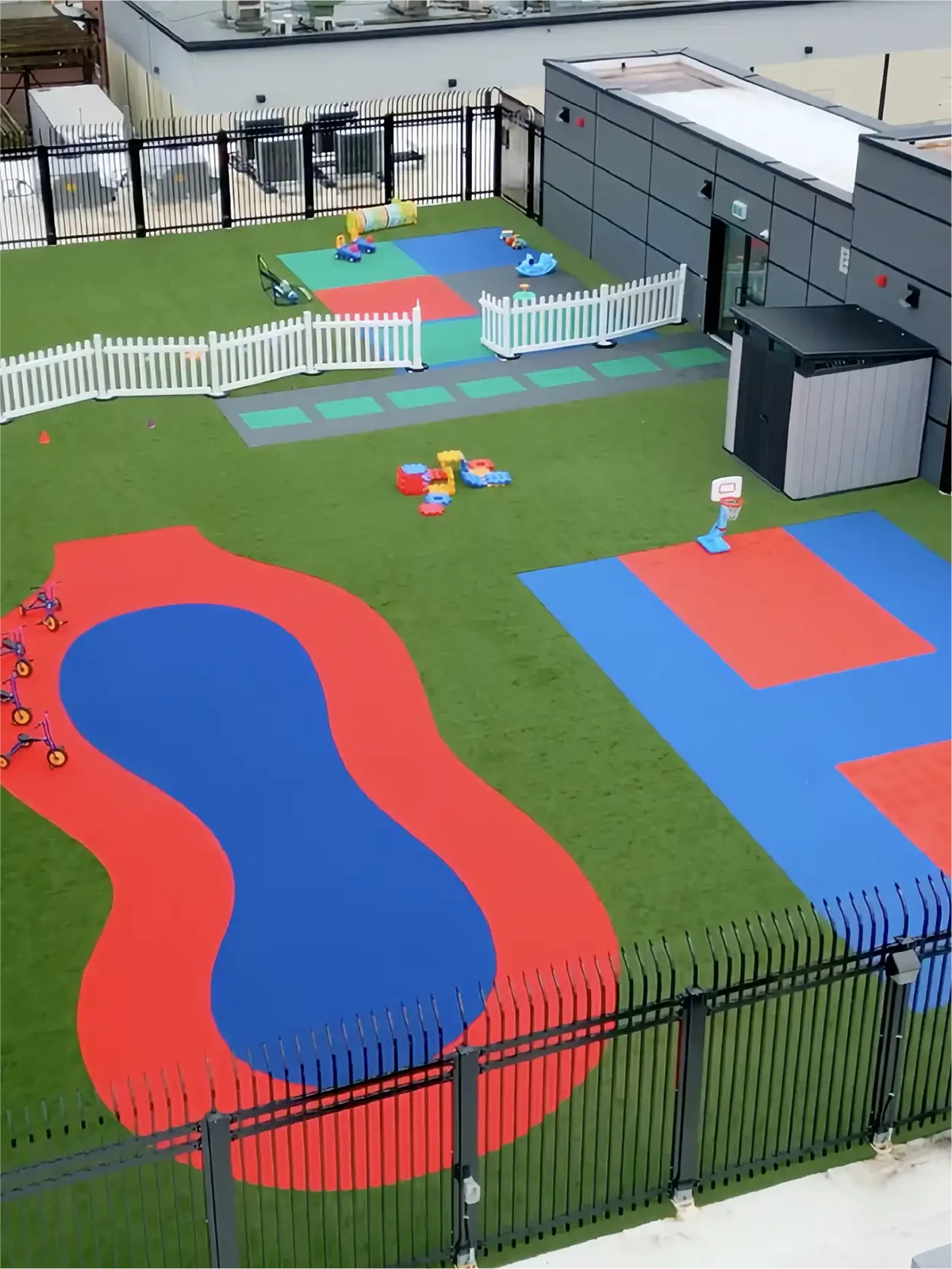 An outdoor playground at early learning centres features artificial grass, red and blue play mats, tricycles, small plastic toys, a mini basketball hoop, a fenced area, and a modern building in the background.