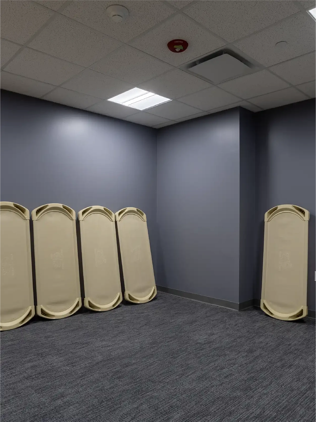 Five beige plastic cots are propped upright against a blue-gray wall in an empty, carpeted room with overhead fluorescent lighting, typical of early learning centres and preschools close to me.