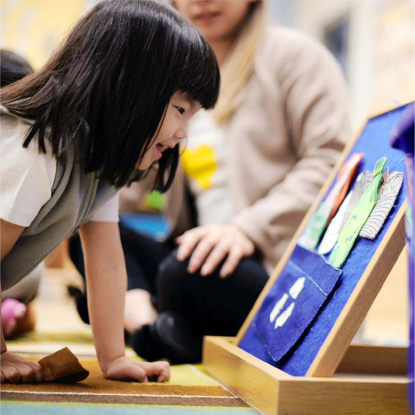 A young child leans in and smiles while looking at a colorful felt board, as an adult from an early childhood care program sits nearby, in a classroom setting.