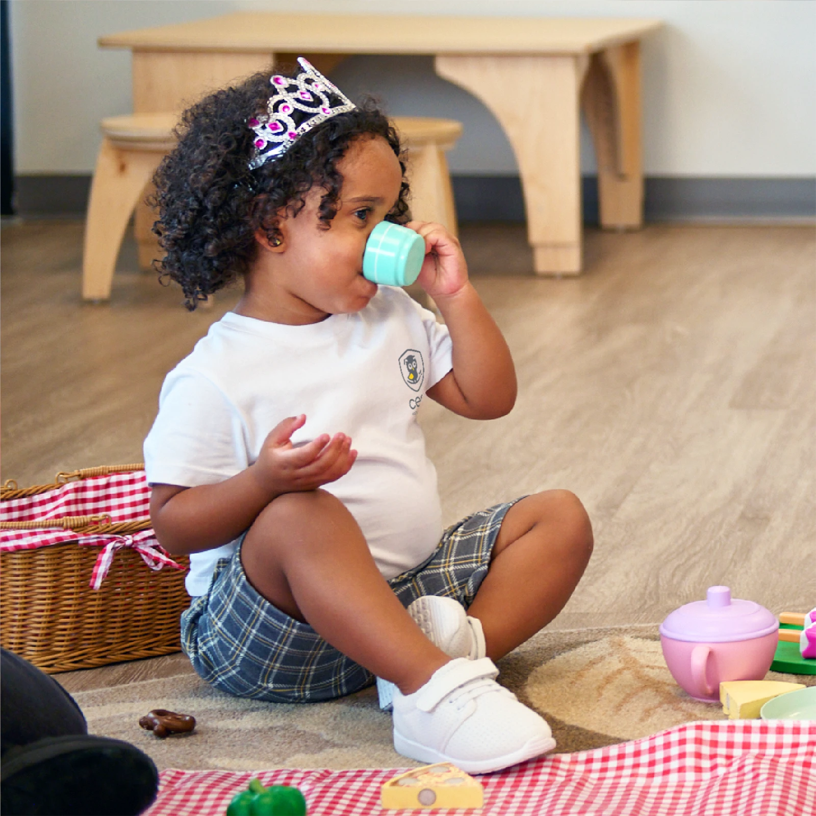 A young child wearing a tiara and plaid shorts sits on the floor, sipping from a toy teacup at a pretend tea party—an imaginative moment often encouraged in early learning centres through engaging play and creative activities.