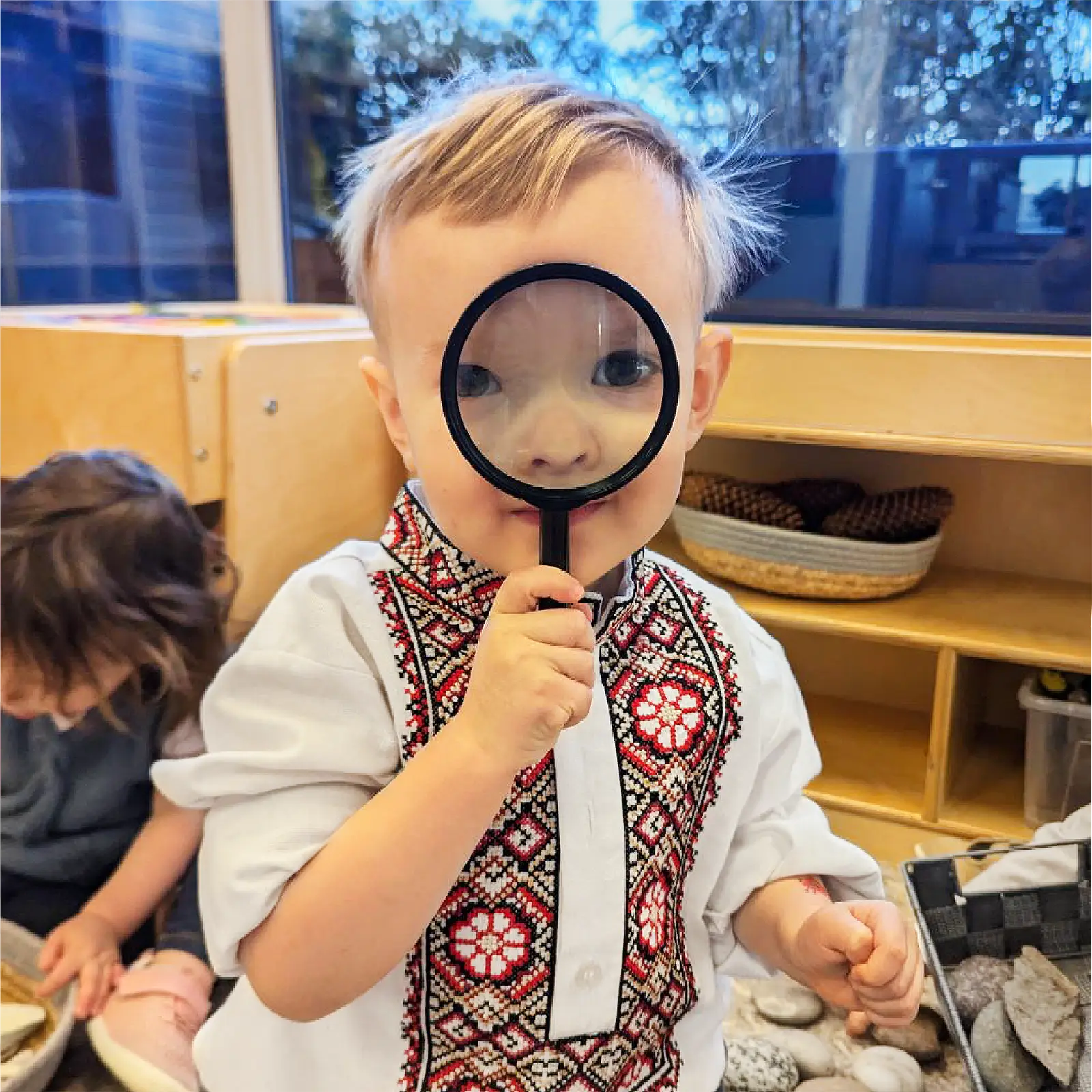 A young child in an embroidered shirt holds a magnifying glass to his face, enlarging one eye. Another child plays in the background of an early learning centre classroom filled with engaging activities.