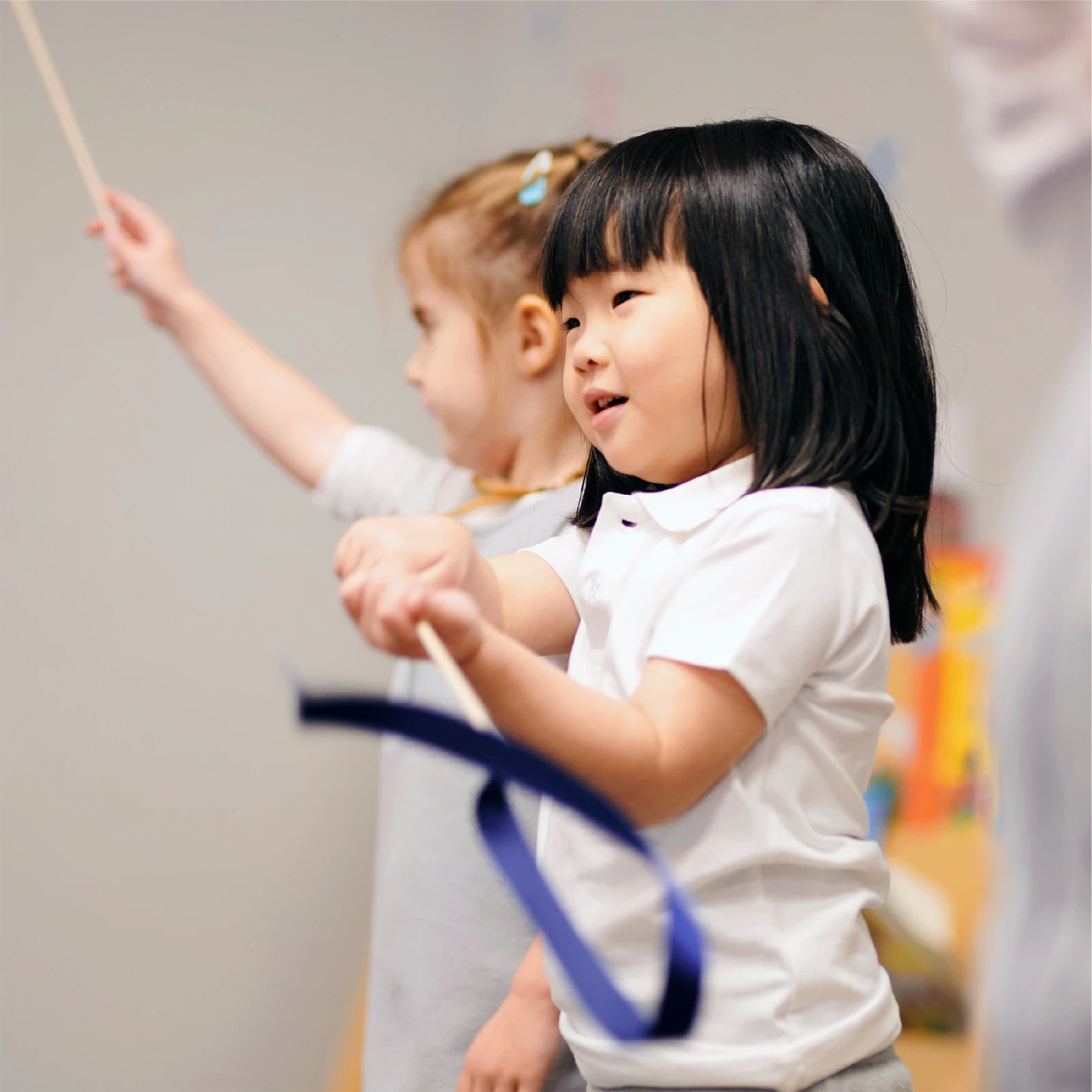 Two young girls smile and wave ribbons in the air during an indoor activity at one of the early learning centres. The girl in the foreground wears a white shirt and holds a blue ribbon, while the girl behind her wears a light gray top.