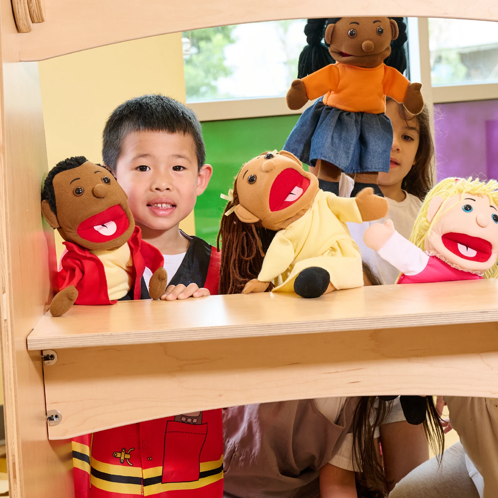 A young child smiles while holding up colorful hand puppets with other children partially visible behind a wooden puppet theater, enjoying creative play at one of the early learning centres known for their diverse and engaging early childhood care program.