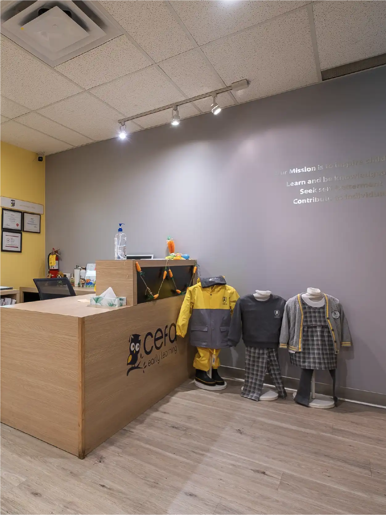 A modern reception area at CEFA Early Learning, one of the leading early learning centres, featuring a wooden desk, children’s uniforms on display, and informational text on a gray wall in a bright and welcoming atmosphere.