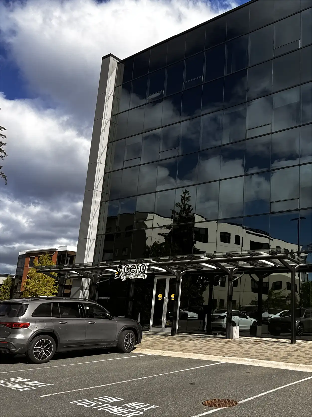 A modern glass office building with reflections of clouds and trees, a parked gray SUV in front, and visible signs for "Celo" above the entrance. Nearby, several early learning centres offer quality early childhood care programs. The sky is partly cloudy.
