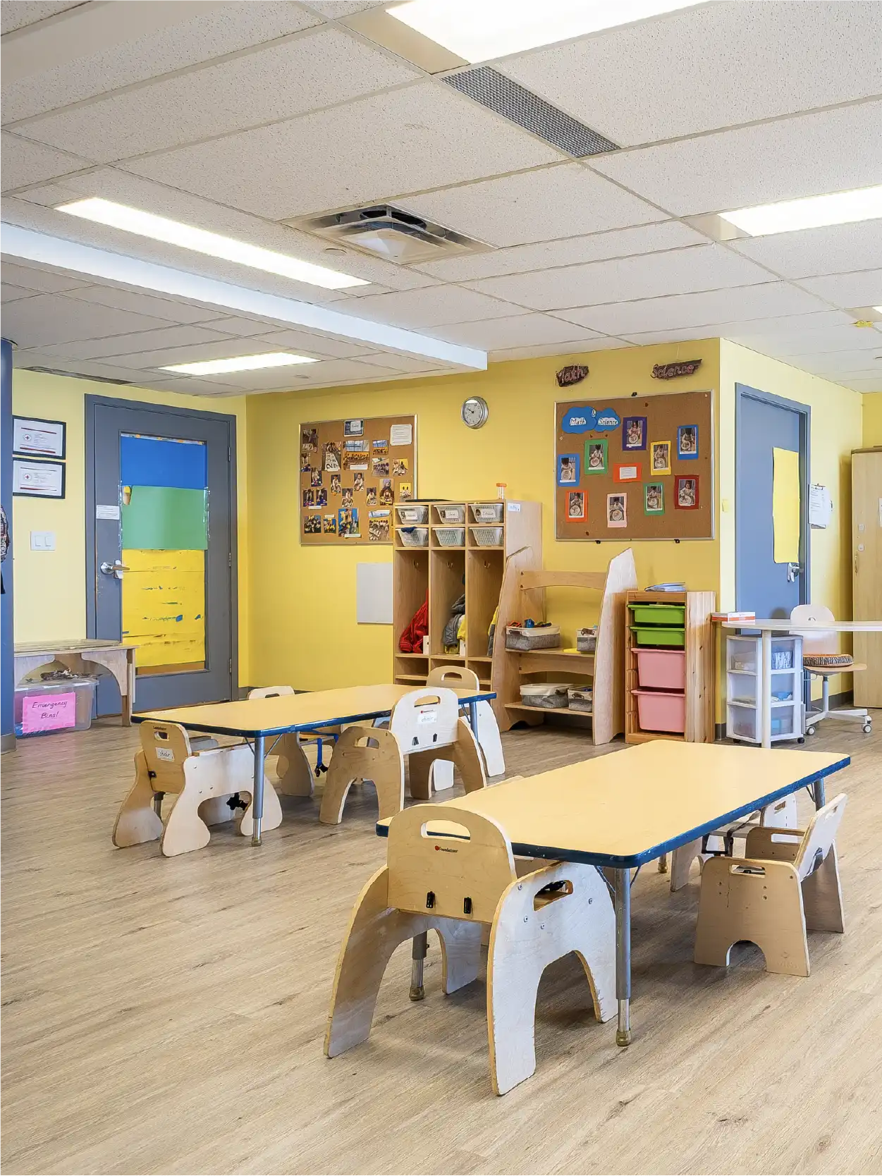 A bright classroom in early learning centres with yellow walls, small wooden tables and chairs, cubbies, colorful storage bins, and children's artwork on bulletin boards. The room has a clean, organized, and welcoming atmosphere.