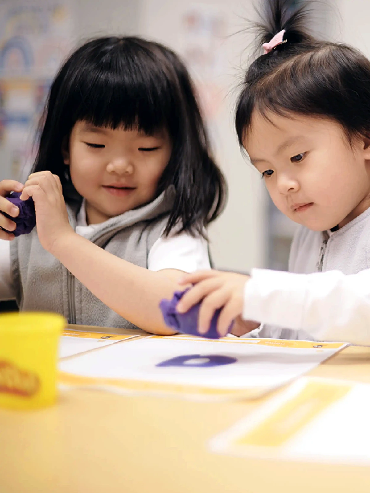 Two young children sit at a table in an early childhood care program, focused on making shapes with purple Play-Doh on sheets of paper. One has short black hair and the other a ponytail; both are fully engaged in their creative activity.