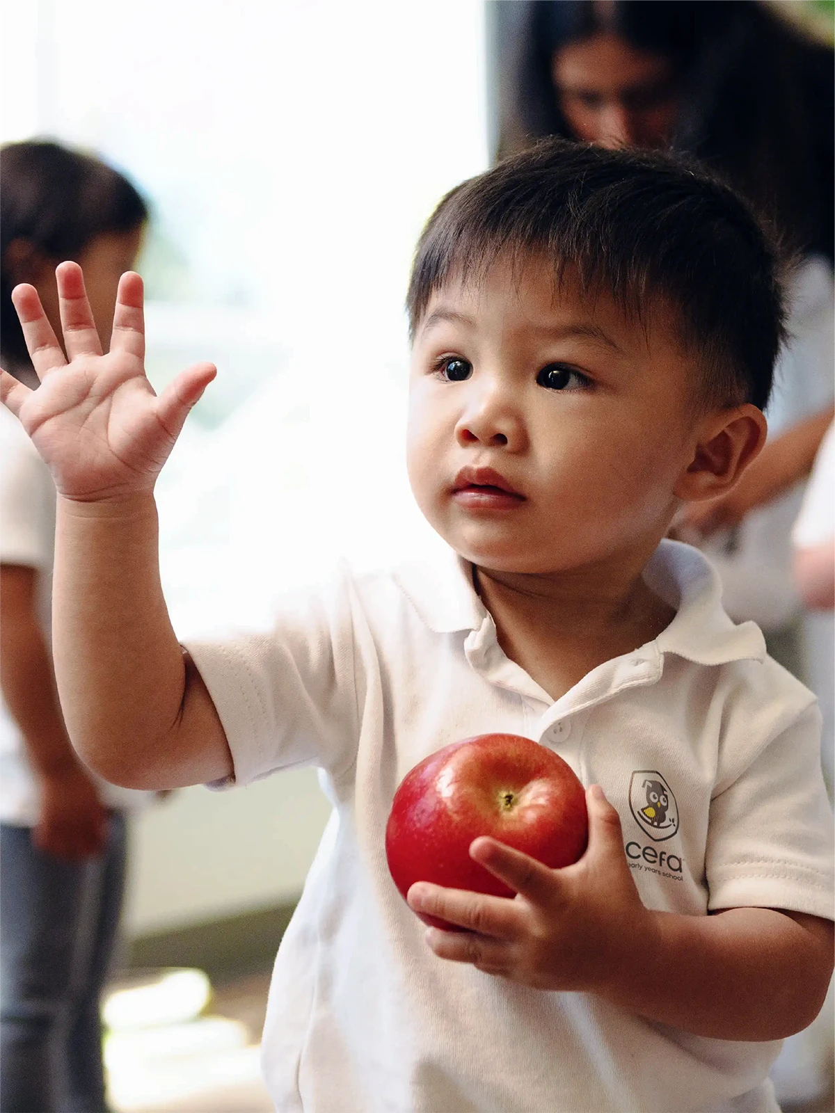 A young child at one of the early learning centres, wearing a white polo shirt, holds a red apple in one hand and raises the other, as if waving or asking a question, in a bright indoor setting.