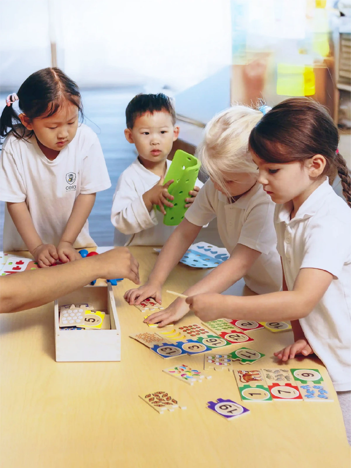 Four young children in white shirts gather around a table, engaging with colorful number and puzzle pieces, while an adult's hand assists them during an educational activity at one of the early learning centres nearby.