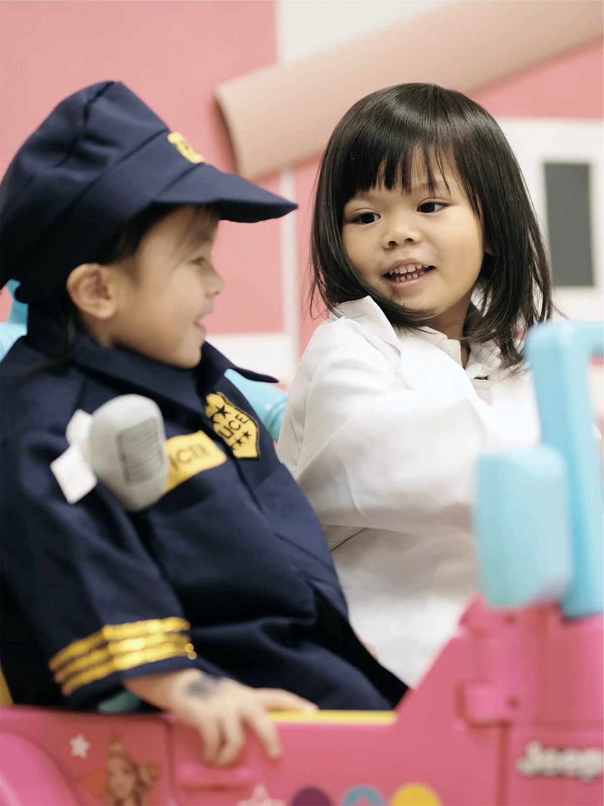 Two young children playing; one dressed as a police officer and the other in a white coat, both smiling in a pink toy car—capturing the joyful spirit found at early learning centres or through an early childhood care program.