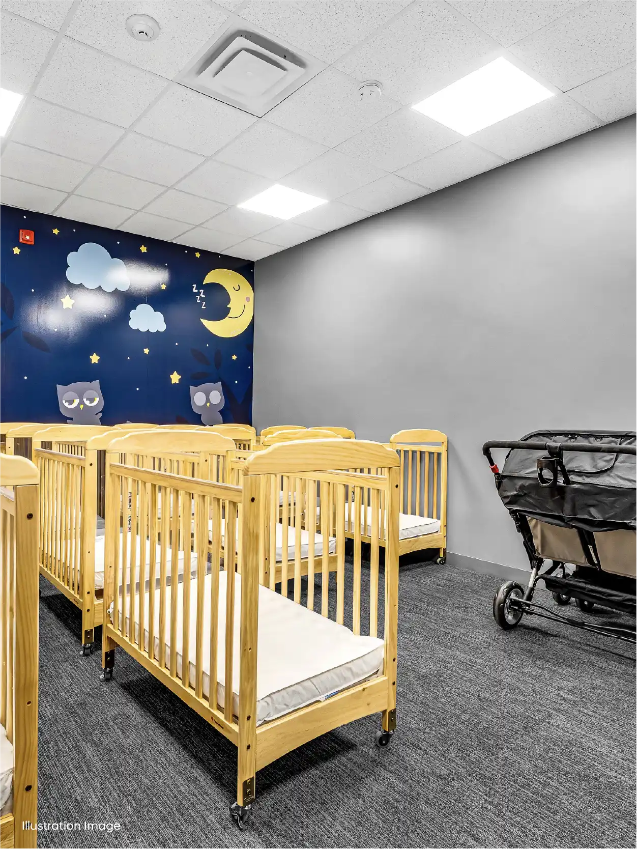 A brightly lit nursery room with several wooden cribs, a black stroller, and a dark blue wall decorated with moon, stars, clouds, and cartoon owls—perfect for an early childhood care program. The gray carpet adds to the simple, clean decor.