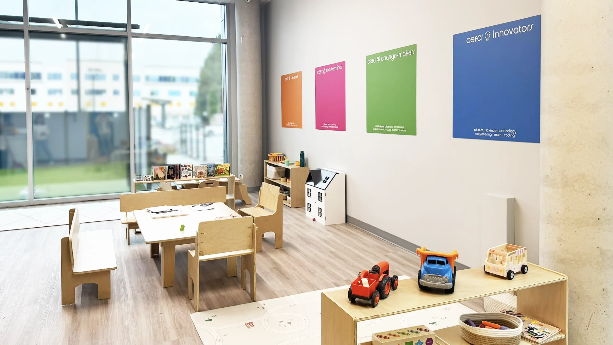 A bright, modern classroom in one of the early learning centres, with wooden tables, chairs, shelves holding toys and books, and colorful educational posters. Large windows let in natural light and show a view of buildings outside.