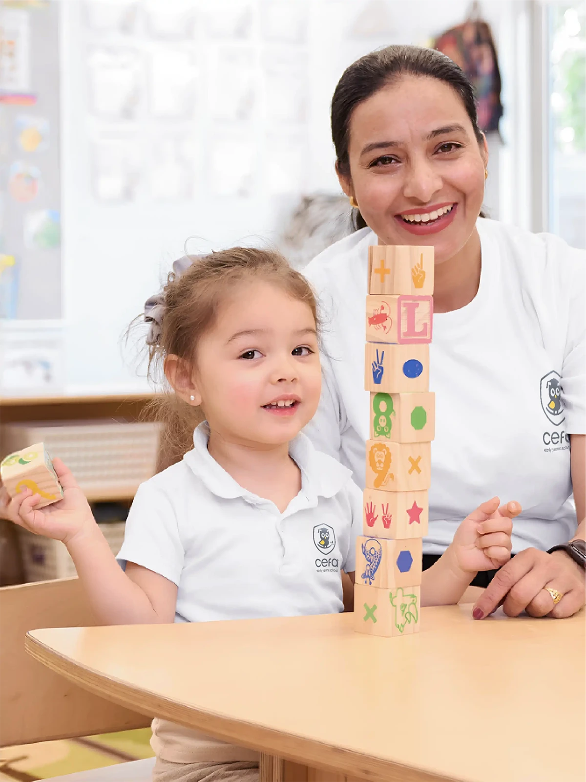 A smiling young girl and an adult woman sit at a table, stacking colorful wooden blocks with symbols and letters—their joy and engagement reflect the welcoming atmosphere found in quality early learning centres.