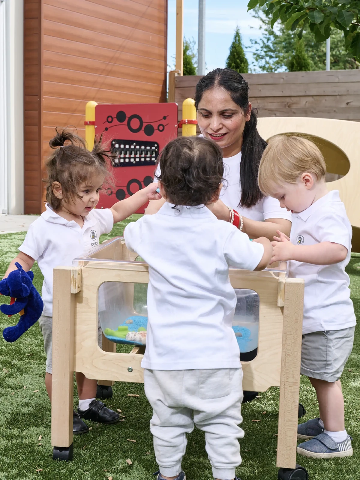 A woman and three toddlers stand around a sensory play table outdoors at an early learning centre. The children are engaged with the toys, and one holds a blue plush toy. They are all wearing white shirts and light-colored pants.