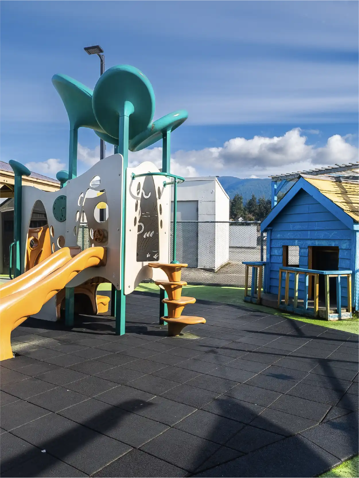 A colorful outdoor playground at an early learning centre features a slide, climbing steps, and a small blue playhouse on a rubber-tiled surface, surrounded by a fence and buildings under a partly cloudy sky.