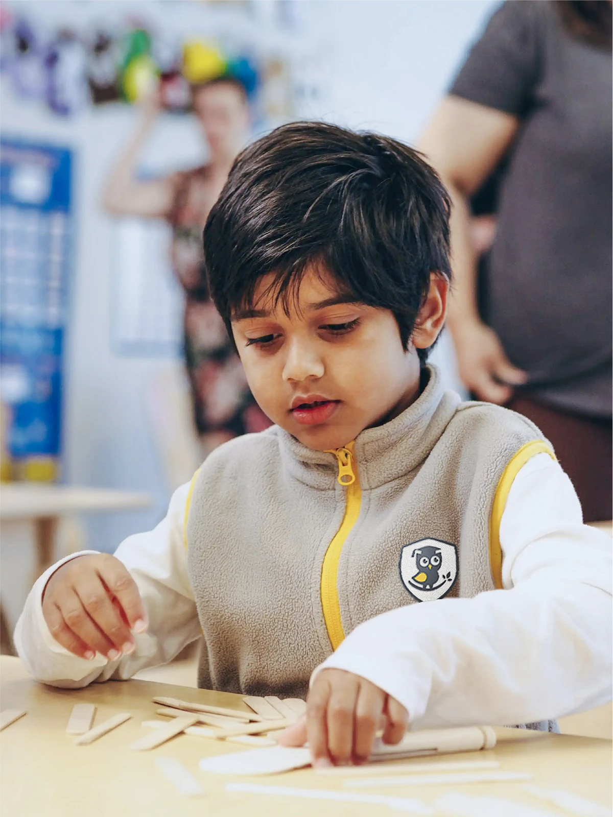 A young child with dark hair, wearing a gray and yellow vest, sits at a table focusing on arranging wooden sticks in an early learning centre classroom, with blurred adults in the background.