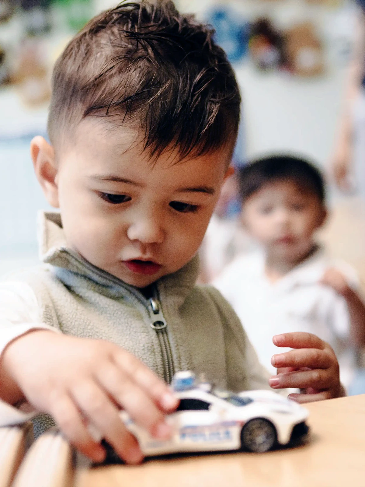 A young child with short brown hair plays with a toy police car on a table at one of the early learning centres, while another child stands blurred in the background.
