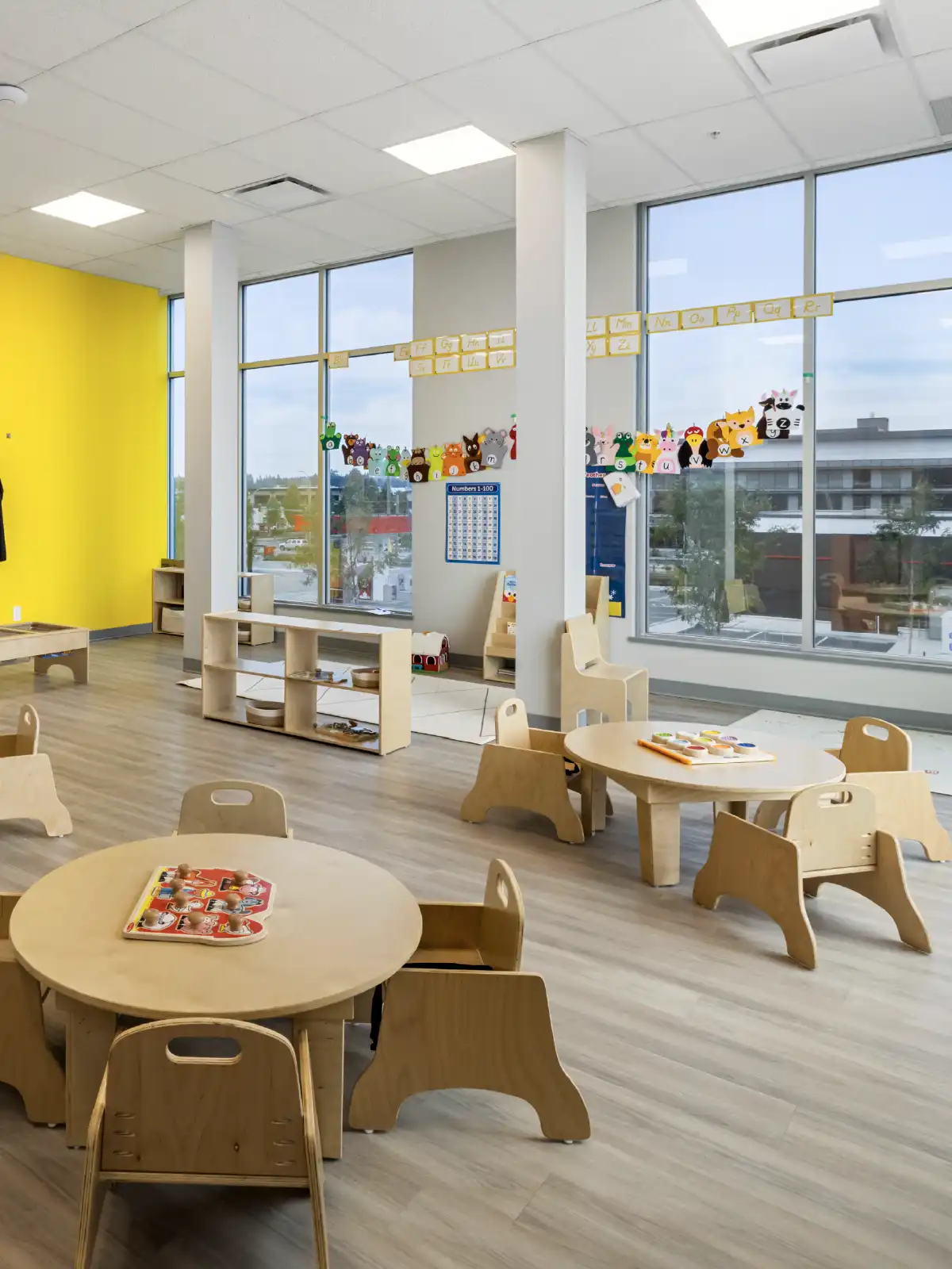 A bright classroom in an early learning centre with round wooden tables, small chairs, toys on shelves, colorful wall decorations, and large windows letting in natural light. Yellow and white walls complement the light wood floor.